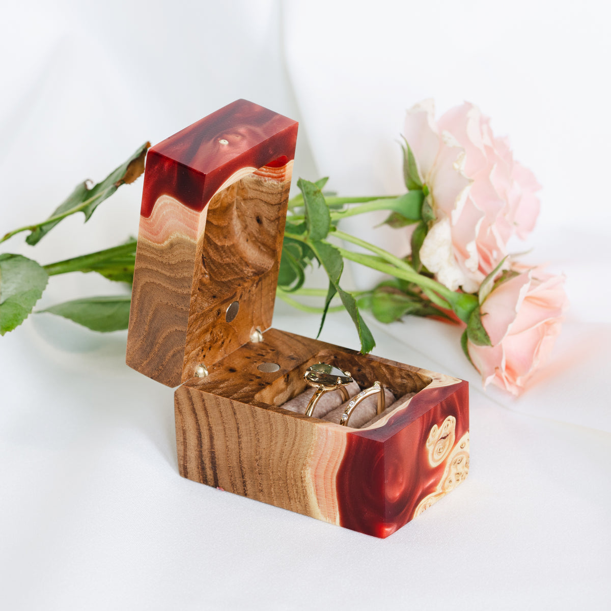Ruby epoxy and elm wood wedding ring box open with two gold rings, pink roses on white satin