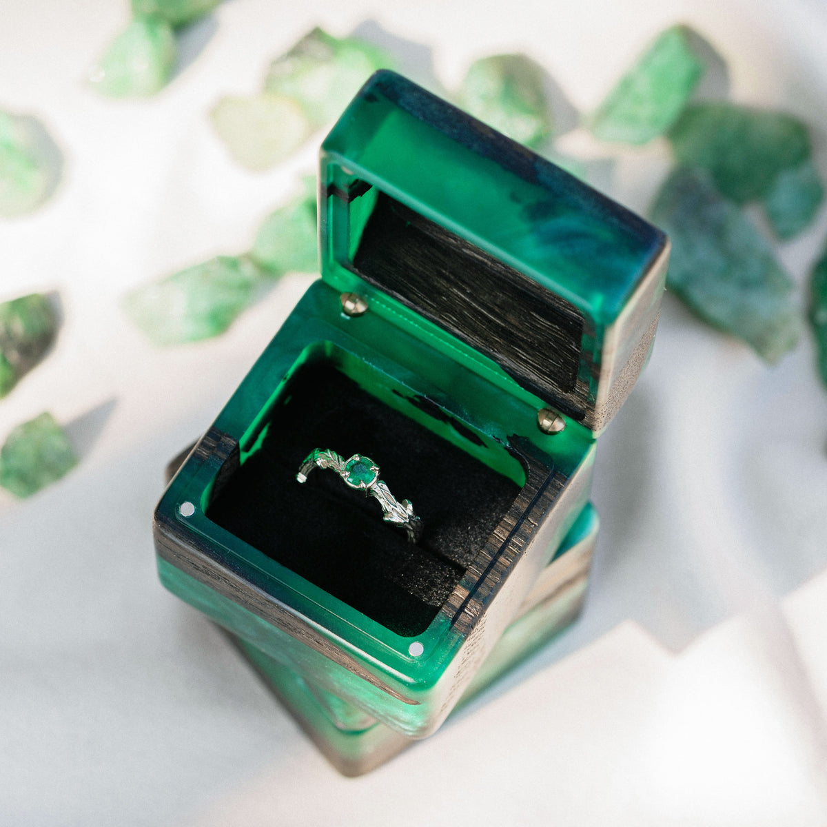 Open green epoxy and bog oak engagement ring box with a gemstone ring, raw emerald stones beside