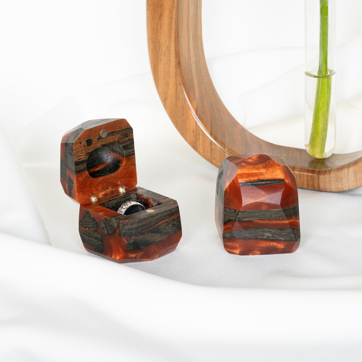 Open and closed amber epoxy ring boxes on white linen by a wooden arch and a glass bud vase