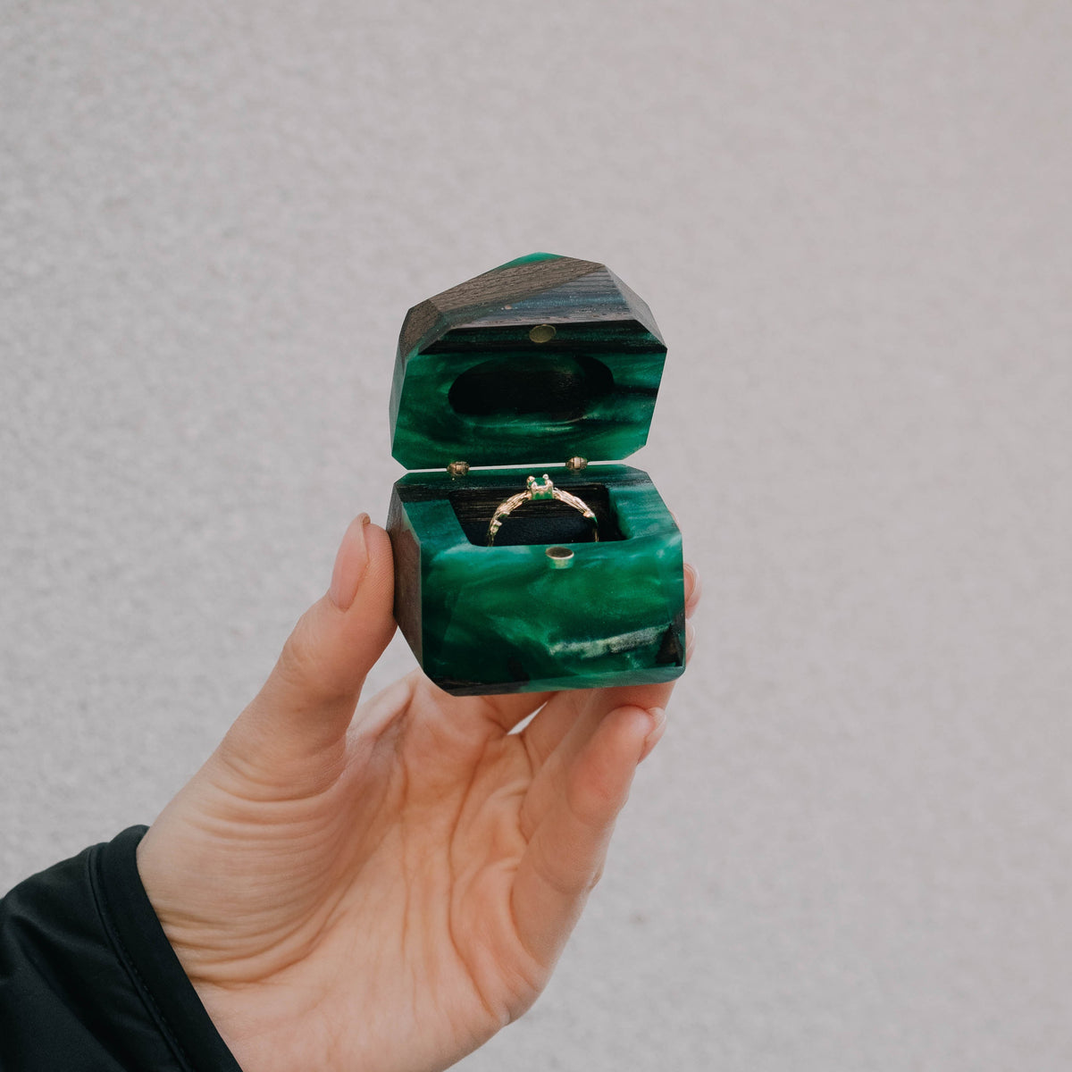 Open green METEORITE box with a gold gemstone ring held up in one hand against a grey wall