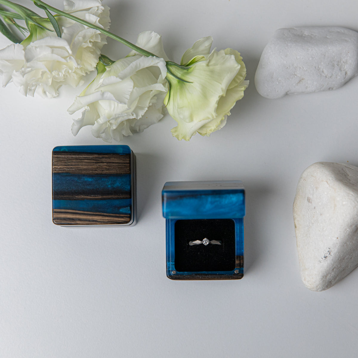 Blue epoxy bog oak ring box open with a diamond ring, flat lay with white stones and flowers